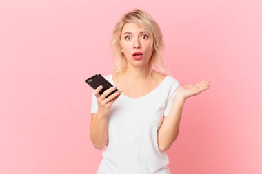 young pretty woman looking surprised and shocked, with jaw dropped holding an object. cell concept