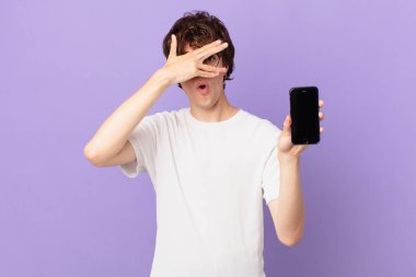 young man looking shocked, scared or terrified, covering face with hand and holding a cell
