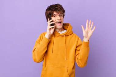 Bir hücreyi tutan genç adam mutlu bir şekilde gülümsüyor, el sallıyor, seni selamlıyor ve selamlıyor.