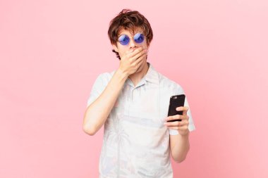 Cep telefonu olan genç bir adam ağzı kapalı ve şok olmuş bir şekilde elleriyle.