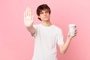 Elinde kahve olan genç adam ciddi görünüyor. Avuç içini gösteriyor. Durdurma hareketi yapıyor.