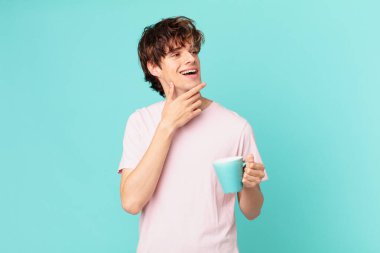 Elinde kahve fincanı olan genç adam mutlu bir şekilde gülümsüyor ve hayal kuruyor ya da şüphe ediyor.