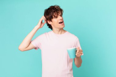 Elinde kahve fincanı olan kafası karışmış, kafası karışmış, kaşınan genç bir adam.