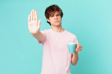 Elinde kahve fincanı olan genç adam ciddi görünüyor. Avuç içini gösteriyor. Durdurma hareketi yapıyor.