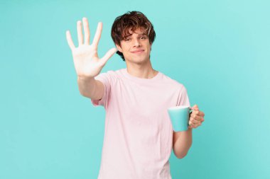 Elinde kahve fincanı olan genç adam gülümsüyor ve arkadaş canlısı görünüyor, beş numarayı gösteriyor.