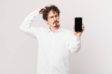 Kafası karışmış, kafası karışmış, kaşınan bir hücreye sahip genç iş adamı.