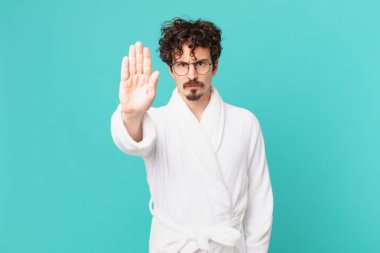 Bornoz giyen genç adam ciddi görünüyor. Avuç içini gösteriyor. Durdurma hareketi yapıyor.