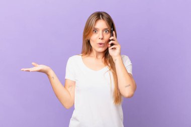 Cep telefonlu genç bir kadın şaşırmış ve şok olmuş görünüyor, çenesi düşmüş bir nesne tutuyor.