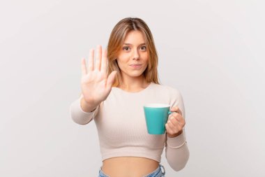Elinde kahve fincanı olan genç bir kadın ciddi görünüyor. Avuç içini gösteriyor. Dur hareketi yapıyor.