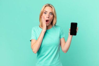 Genç sarışın kadın şok olmuş, korkmuş ve elinde akıllı bir telefon tutuyor.