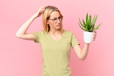 Genç sarışın kadın şaşkın ve kafası karışmış hissediyor. Kafasını kaşıyor ve elinde kaktüs tutuyor.