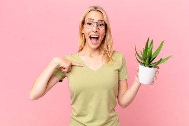 Genç sarışın kadın kendini mutlu hissediyor ve heyecanlı ve elinde bir kaktüsle kendini işaret ediyor.