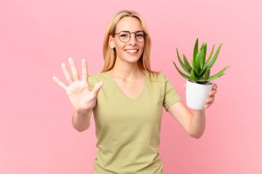Genç sarışın kadın gülümsüyor ve arkadaş canlısı görünüyor, beş numarayı gösteriyor ve elinde bir kaktüs tutuyor.