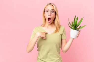 Genç sarışın kadın şok olmuş ve şaşırmış görünüyor ağzı sonuna kadar açık, kendini gösteriyor ve elinde bir kaktüs tutuyor.
