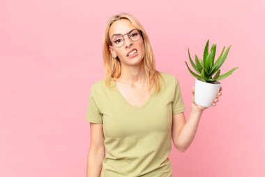 Genç sarışın kadın şaşkın, kafası karışmış ve elinde bir kaktüs tutuyor.