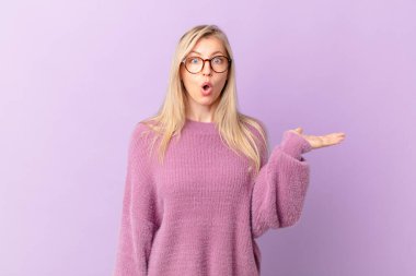 Genç sarışın kadın şaşırmış ve şok olmuş görünüyor, çenesi düşmüş, elinde bir nesne tutuyor.