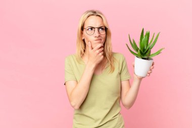 Genç sarışın kadın düşünüyor, kuşkulu ve şaşkın hissediyor ve elinde bir kaktüs tutuyor.