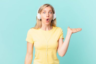 young blonde woman looking surprised and shocked, with jaw dropped holding an object and listening music.