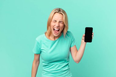 Genç sarışın kadın agresif bir şekilde bağırıyor, kızgın görünüyor ve elinde akıllı bir telefon var.