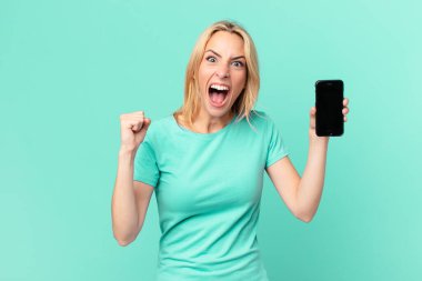 Genç sarışın kadın sinirli bir ifadeyle bağırıyor ve elinde akıllı bir telefon tutuyor.