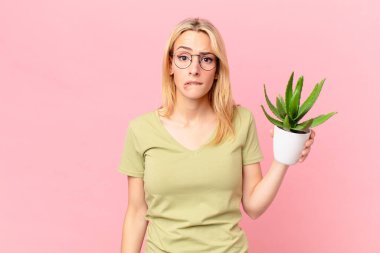 Şaşkın, kafası karışmış ve elinde kaktüs tutan genç sarışın kadın.