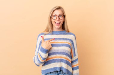 Genç sarışın kadın şok olmuş ve şaşırmış görünüyor, ağzı açık, kendini işaret ediyor.