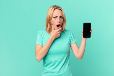 Genç sarışın kadın, ağzı ve gözleri sonuna kadar açık, eli çenesinde ve elinde akıllı bir telefonla.