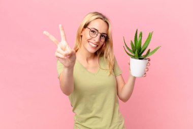 Genç sarışın kadın gülümsüyor ve mutlu görünüyor, zafer ya da barış işareti yapıyor ve elinde bir kaktüs tutuyor.