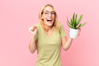 Genç sarışın kadın şokta, gülüyor, başarıyı kutluyor ve elinde bir kaktüs tutuyor.
