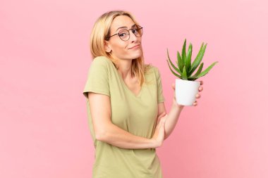 Genç sarışın kadın omuz silkiyor, kafası karışık ve kararsız ve elinde bir kaktüs tutuyor.