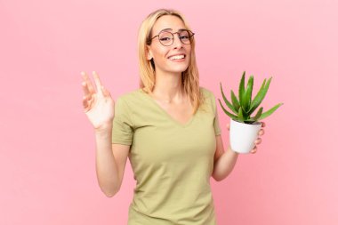 Genç sarışın kadın mutlu bir şekilde gülümsüyor, el sallıyor, seni selamlıyor ve bir kaktüs tutuyor.
