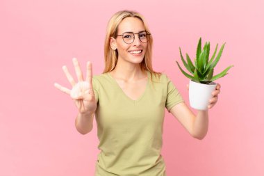 Genç sarışın kadın gülümsüyor ve arkadaş canlısı görünüyor, dört numarayı gösteriyor ve elinde bir kaktüs tutuyor.
