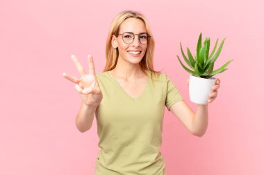 Genç sarışın kadın gülümsüyor ve arkadaş canlısı görünüyor, üç numarayı gösteriyor ve elinde bir kaktüs tutuyor.