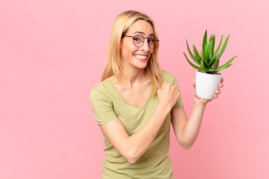 Genç sarışın kadın kendini mutlu hissediyor ve meydan okumayla yüzleşiyor ya da kutluyor ve kaktüs tutuyor.