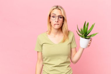 Şaşkın, kafası karışmış ve elinde kaktüs tutan genç sarışın kadın.