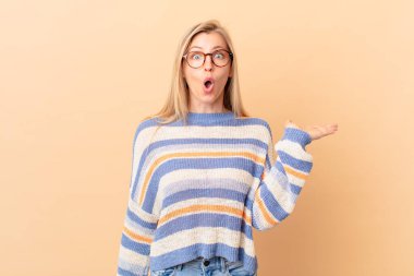 Genç sarışın kadın şaşırmış ve şok olmuş görünüyor, çenesi düşmüş, elinde bir nesne tutuyor.