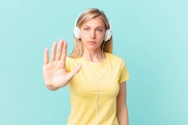 young blonde woman looking serious showing open palm making stop gesture and listening music.