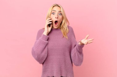 Genç sarışın kadın şok olmuş, şaşırmış ve akıllı telefonuyla konuşuyor.