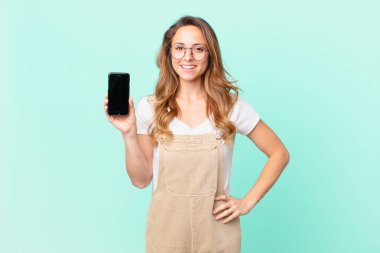 Güzel bir kadın mutlu bir şekilde gülümsüyor, eli kalçasında, kendinden emin ve akıllı bir telefon tutuyor.
