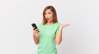 Güzel bir kadın şaşırmış ve şok olmuş görünüyor, çenesi düşmüş, elinde bir nesne tutuyor ve akıllı telefon kullanıyor.