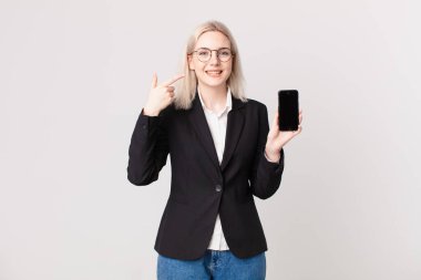 Sarışın, güzel bir kadın. Kendine güvenerek gülümsüyor. Geniş bir gülümseme ve cep telefonu tutuyor.