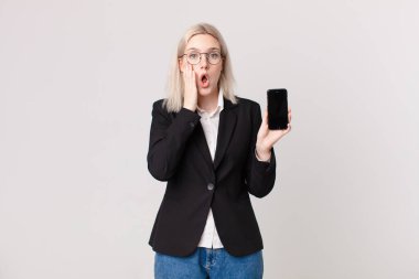 Sarışın, güzel bir kadın şok olmuş, korkmuş ve elinde cep telefonu tutuyor.