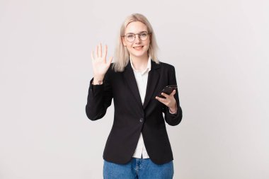 Sarışın, güzel bir kadın gülümsüyor ve arkadaş canlısı görünüyor. Beş numarayı gösteriyor ve elinde bir cep telefonu var.