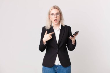 Sarışın, güzel bir kadın ağzı açık, şok olmuş ve şaşırmış görünüyor. Kendini gösteriyor ve elinde cep telefonu var.