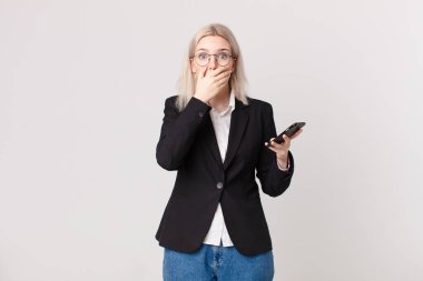 Sarışın, güzel bir kadın elleri ve elleri ile ağzını kapatıyor ve elinde cep telefonu var.