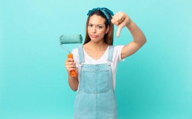 young hispanic woman feeling cross,showing thumbs down and painting a wall