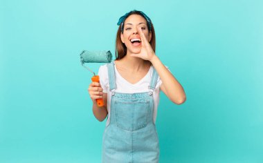 young hispanic woman feeling happy,giving a big shout out with hands next to mouth and painting a wall