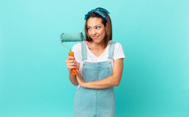 young hispanic woman shrugging, feeling confused and uncertain and painting a wall