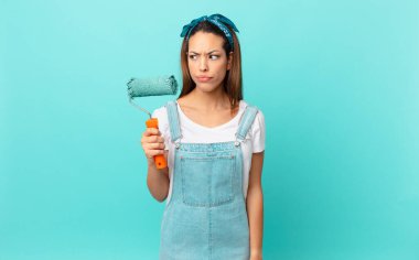 young hispanic woman feeling sad, upset or angry and looking to the side and painting a wall