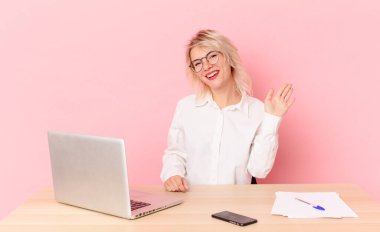 Sarışın, güzel, genç ve güzel bir kadın mutlu bir şekilde gülümsüyor, el sallıyor, sizi selamlıyor. çalışma alanı masa konsepti